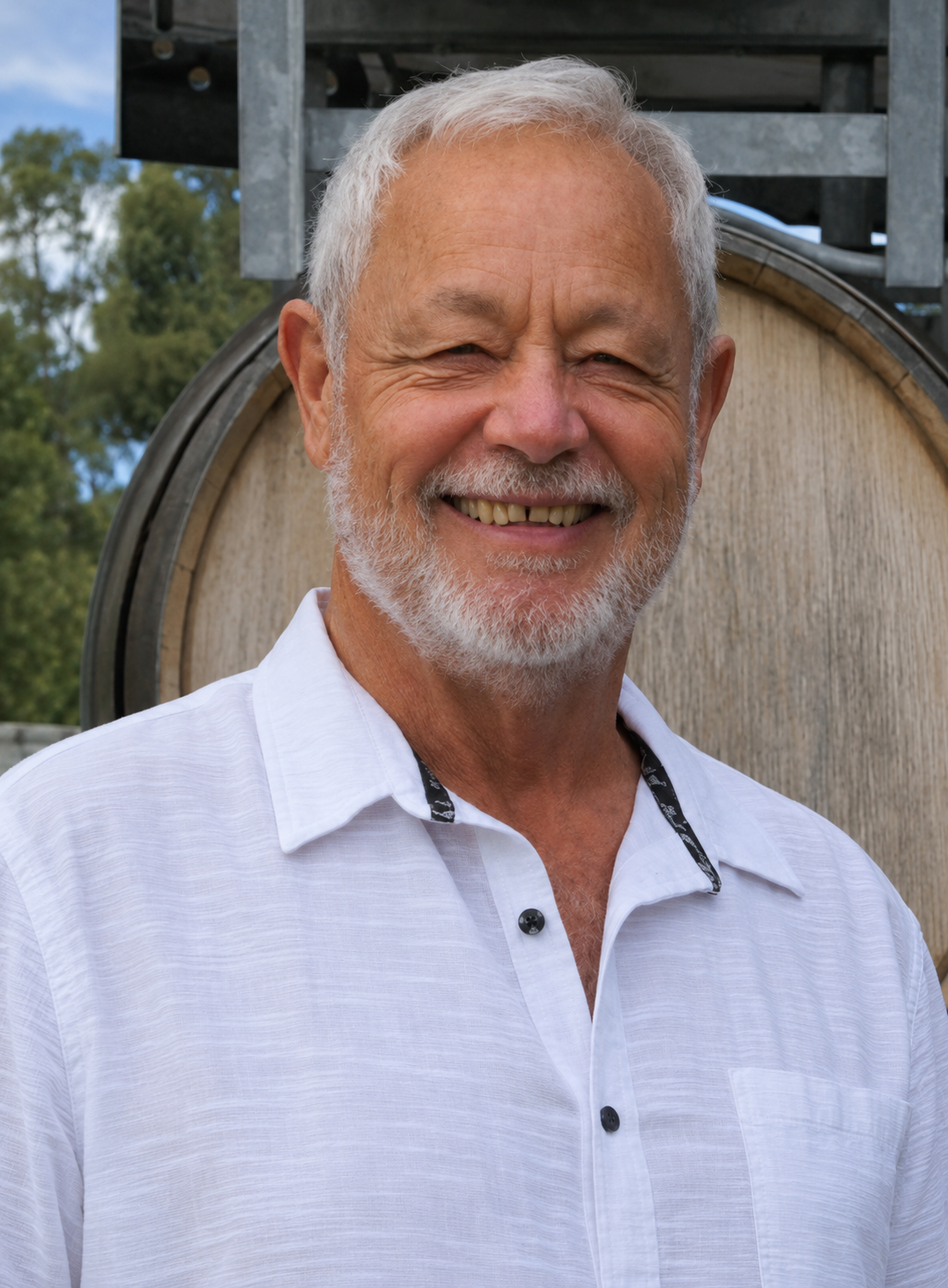 Man with gray hair and beard wearing a white shirt standing in front of a wooden barrel.